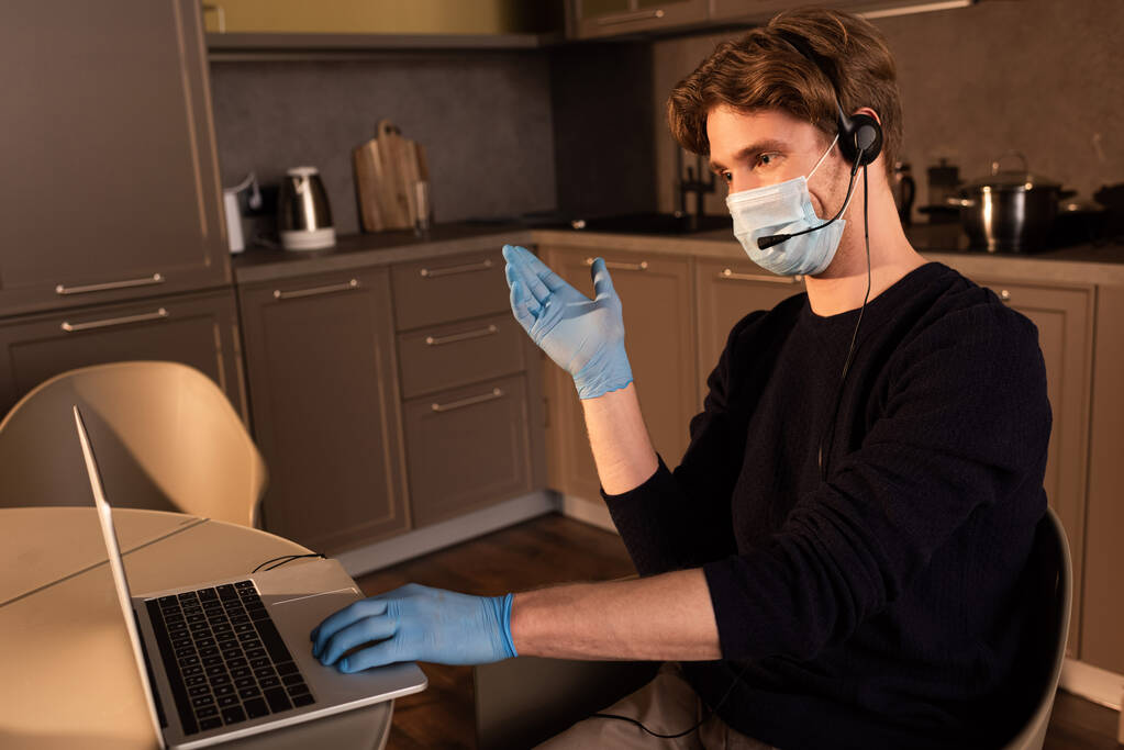 Telepracownik w masce medycznej i rękawicach lateksowych wskazując ręką podczas korzystania z zestawu słuchawkowego i laptopa w kuchni  - Zdjęcie, obraz