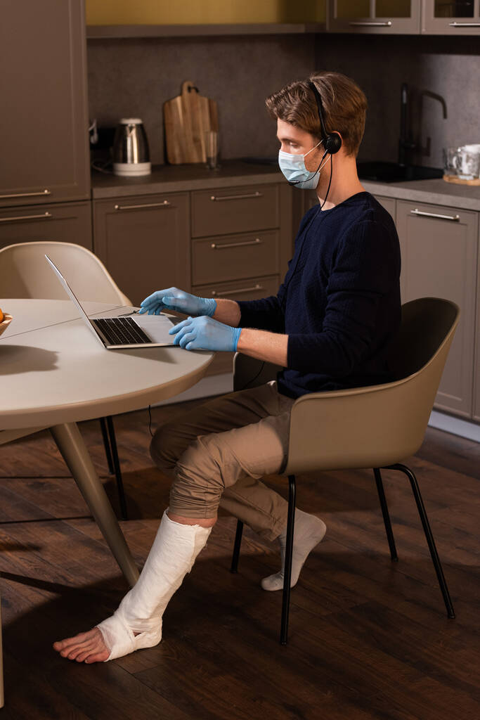Vista laterale dell'uomo disabile in maschera medica e guanti in lattice che lavorano con laptop e auricolare sul tavolo della cucina
  - Foto, immagini