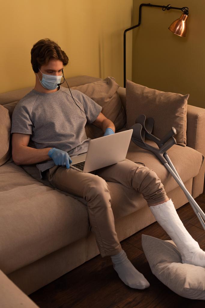 Behinderter in medizinischer Maske arbeitet mit Headset und Laptop in der Nähe von Krücken auf dem Sofa  - Foto, Bild