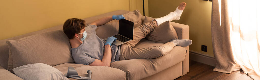 Панорамная концепция фрилансера в медицинской маске со сломанной ногой, работающего на ноутбуке дома
  - Фото, изображение