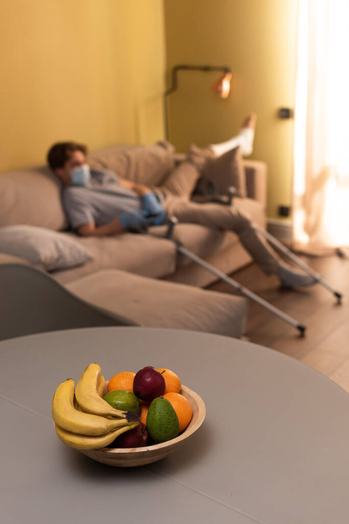 Selektiver Fokus von Früchten auf Tisch und Mann mit gebrochenem Bein hält Fernbedienung auf Couch zu Hause  - Foto, Bild