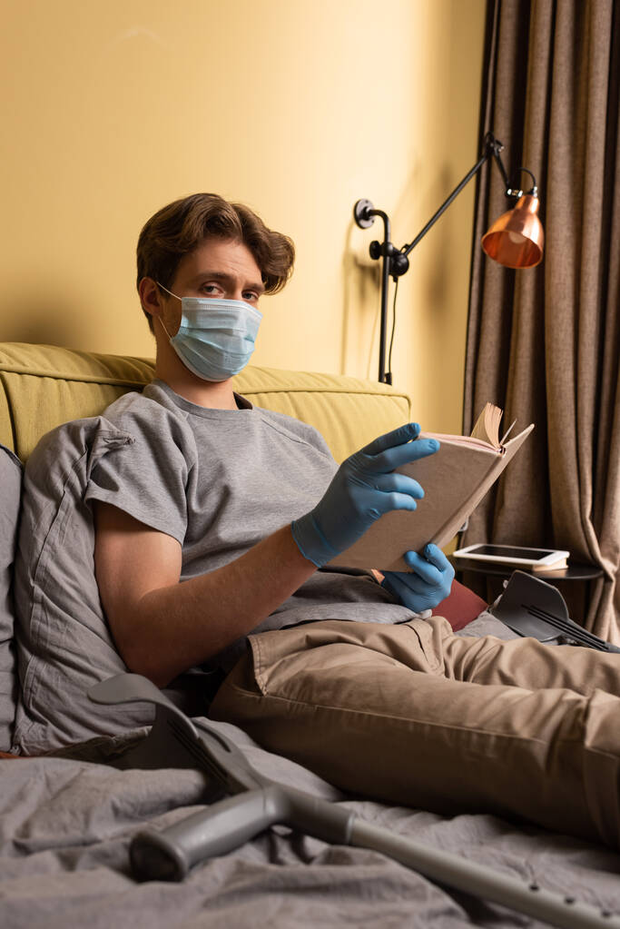 Enfoque selectivo del hombre discapacitado en máscara médica y guantes de látex mirando a la cámara mientras sostiene libro cerca de muletas en la cama
  - Foto, imagen