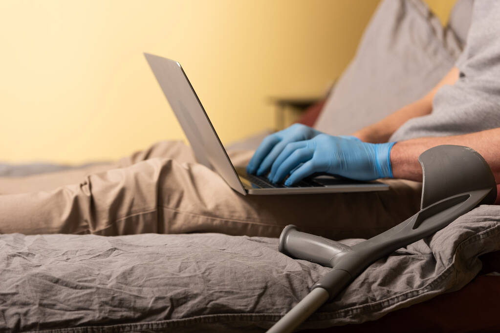 ベッドの上でラップトップを使用してラテックス手袋で障害者の近くの松葉杖の作物ビュー  - 写真・画像