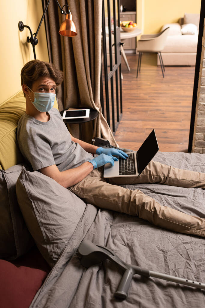 Hombre con máscara médica mirando a la cámara mientras usa el portátil cerca de muletas en la cama
  - Foto, imagen