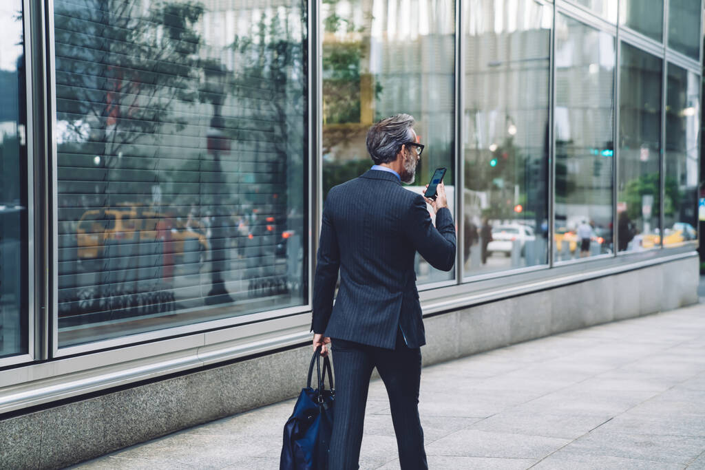 Rückenansicht eines erfolgreichen Mannes mittleren Alters im eleganten dunklen Businessanzug, der mit Smartphone und Tasche die Straße in New York hinunterläuft - Foto, Bild