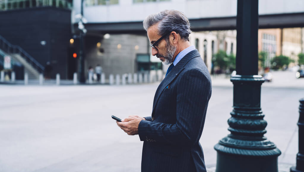 ニューヨークの列の横にある歩道に立ってスマートフォンをタップ成熟したファッショナブルなビジネスマンの側面図ぼやけた背景に - 写真・画像
