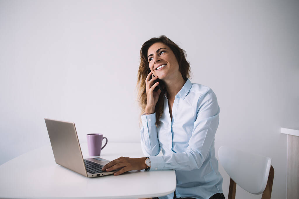 Resmi tişörtlü yetişkin bir kadın telefonda kahve ve dizüstü bilgisayarla oturuyor ve başka tarafa bakıyor. - Fotoğraf, Görsel