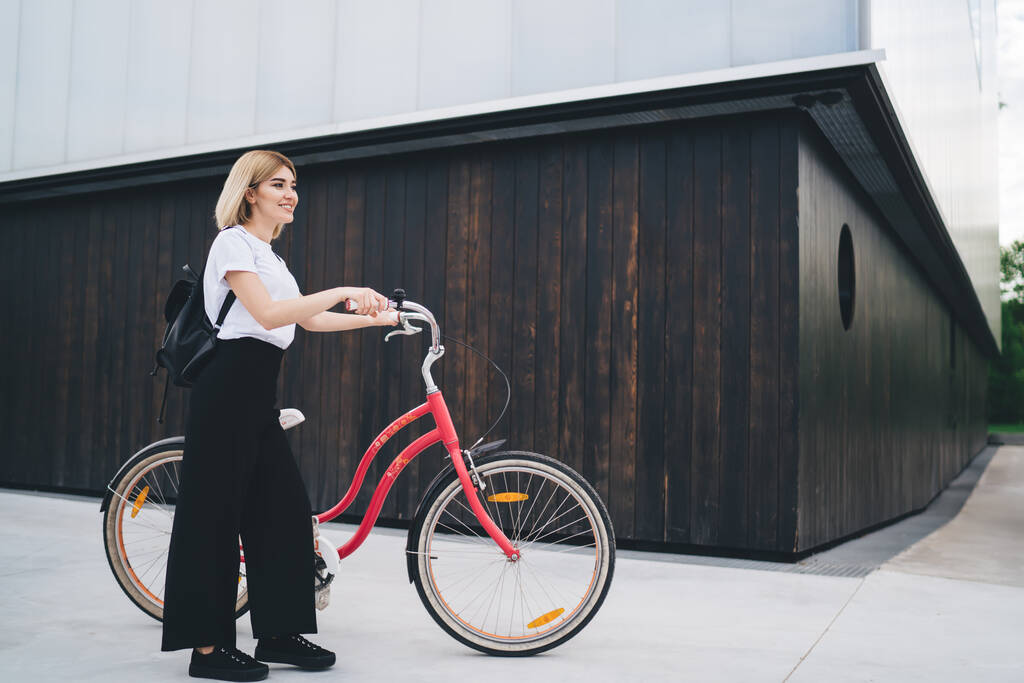 Side view of cheerful young modern blond woman in casual outfit and with black backpack standing with bicycle against street building looking away  - Photo, Image