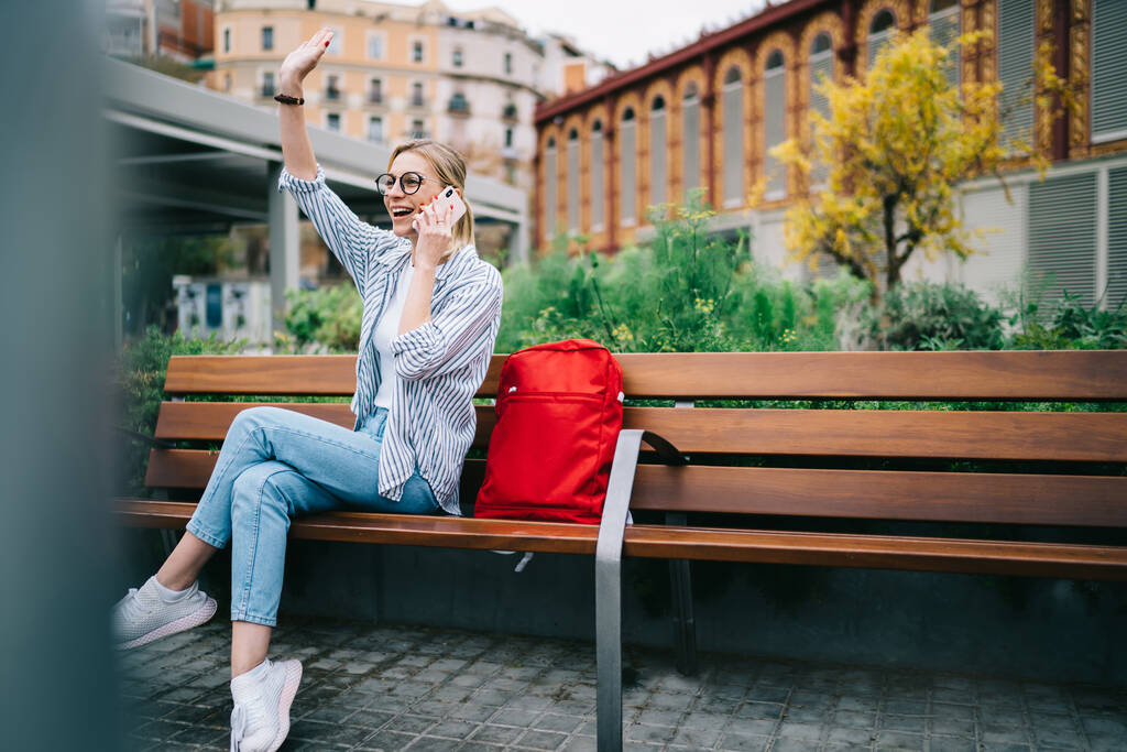 Jovencita alegre en ropa casual sentada cerca de la mochila roja en el banco de madera y saludando con la mano mientras habla en el teléfono inteligente en el parque
 - Foto, imagen