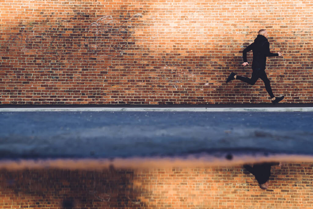 Oldalnézet sportos rövid hajú fiatalember fekete sportruházat és tornacipő kocogás keskeny gyalogúton mentén varrat nélküli narancssárga tégla fal szabadban - Fotó, kép