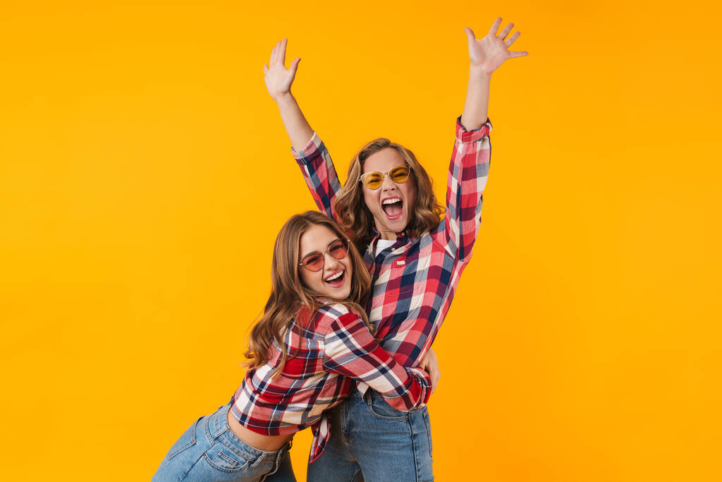 Imagen de dos chicas hermosas jóvenes con camisas a cuadros sonriendo y divirtiéndose aisladas sobre fondo amarillo
 - Foto, imagen