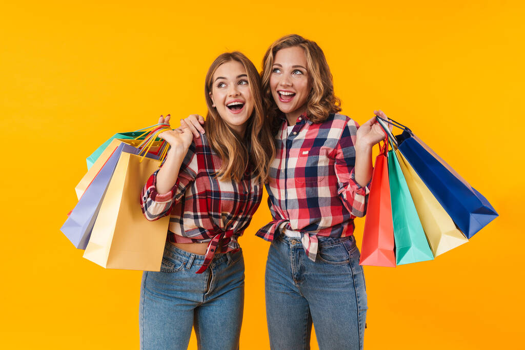 Image of two young beautiful girls wearing plaid shirts smiling and holding colorful shopping bags isolated over yellow background - Photo, Image