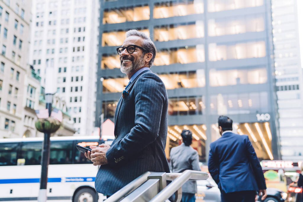 Wesoły biznesmen w średnim wieku w eleganckim garniturze i okularach ze smartfonem opierającym się na poręczy schodów w centrum Nowego Jorku - Zdjęcie, obraz