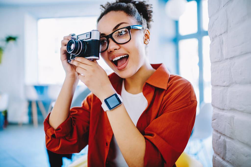 Verrast vrouwelijke amateur fotograaf in optische bril maken van foto via vintage camera, donkere huid verbaasd vrouw met expressie op gezicht nemen van foto 's met behulp van ouderwetse apparatuur binnen - Foto, afbeelding