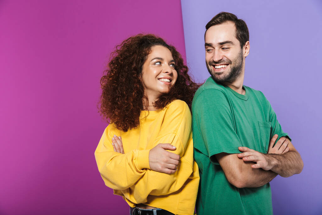 Портрет брюнетки кавказской пары мужчина и женщина в красочной одежде улыбаются друг другу стоя со скрещенными руками на фиолетовом фоне
 - Фото, изображение