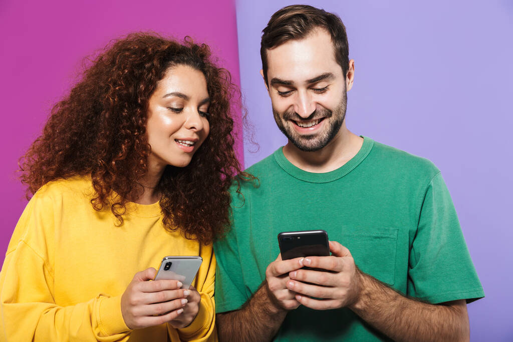 Portret van een prachtig blank echtpaar man en vrouw in kleurrijke kleding glimlachen en houden mobiele telefoons bij elkaar geïsoleerd over violette achtergrond - Foto, afbeelding