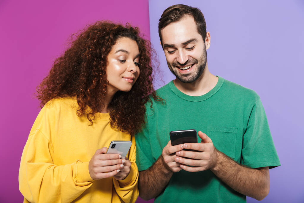 Retrato de alegre pareja caucásica hombre y mujer en ropa colorida sonriendo y sosteniendo los teléfonos celulares juntos aislados sobre fondo violeta
 - Foto, Imagen