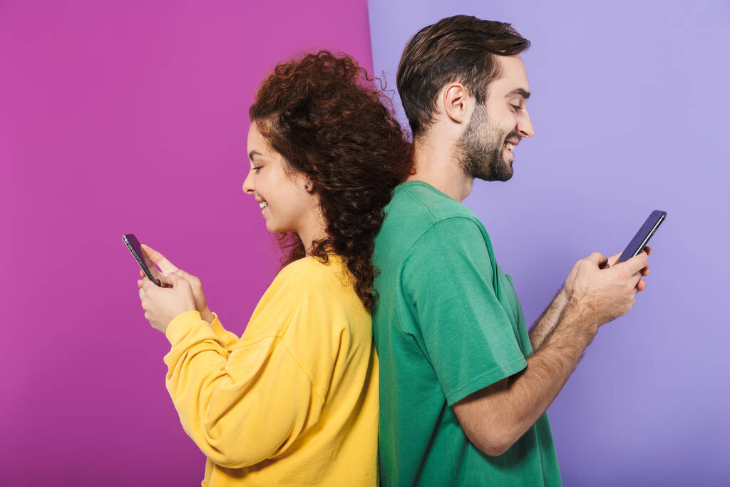 Πορτρέτο του ικανοποιημένου καυκάσιου ζευγαριού με πολύχρωμα ρούχα χαμογελώντας και κρατώντας κινητά τηλέφωνα που στέκονται πλάτη με πλάτη απομονωμένα σε βιολετί φόντο - Φωτογραφία, εικόνα