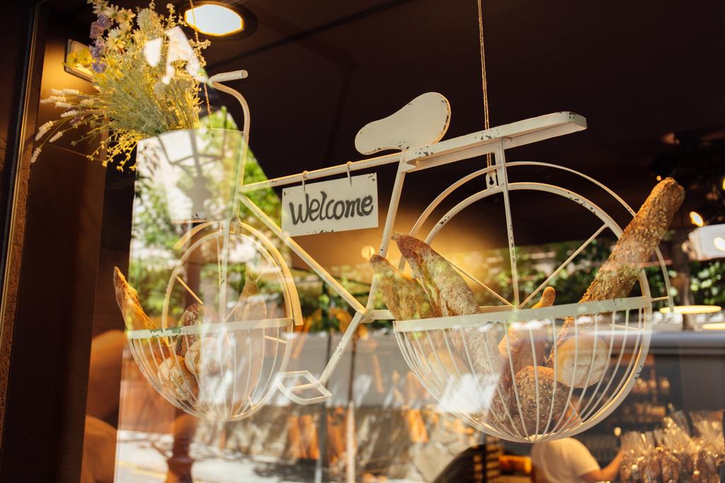 Bicicletta decorativa con fiori, pasticceria e cartolina con scritte di benvenuto in vetrina di panetteria
   - Foto, immagini