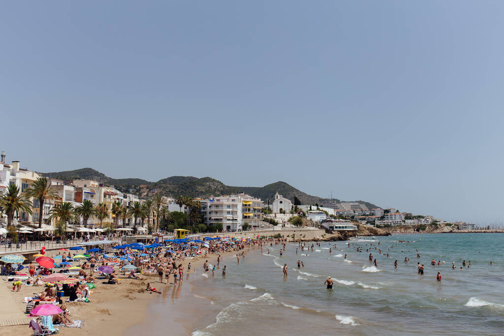 CATALONIA, ΙΣΠΑΝΙΑ - 30 ΑΠΡΙΛΙΟΥ 2020: Άνθρωποι αναπαύονται σε αμμώδη παραλία και κολυμπούν στη θάλασσα κοντά σε κτίρια και φοίνικες στην ακτή - Φωτογραφία, εικόνα