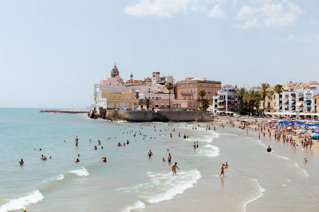 Каталония, Испания - 30 апреля 2020 года: люди плавают в море рядом со зданиями и пальмами на пляже
 - Фото, изображение