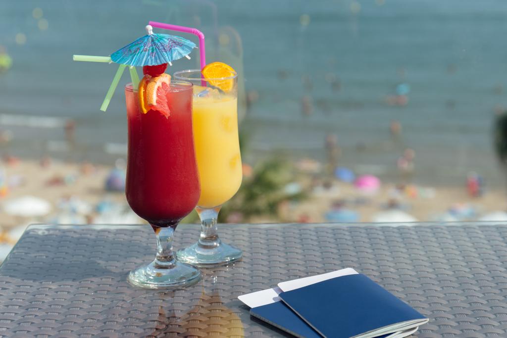 Copas de cócteles y pasaportes con billetes de avión en la mesa con la playa al fondo
 - Foto, Imagen