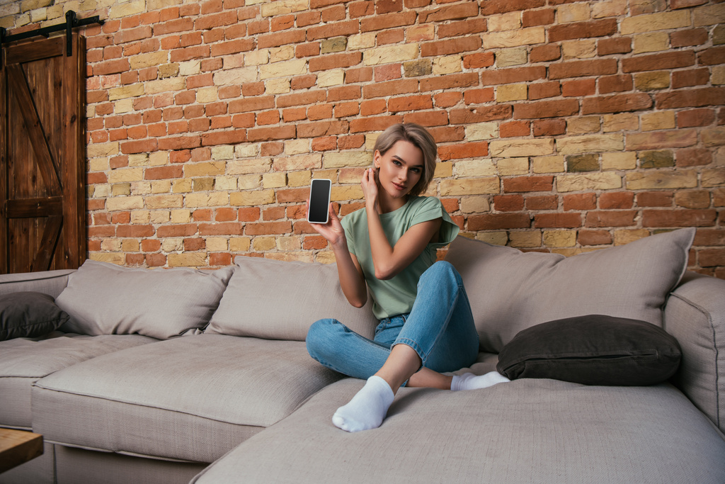 όμορφη νεαρή γυναίκα δείχνει smartphone με κενή οθόνη, ενώ κάθεται στον καναπέ - Φωτογραφία, εικόνα