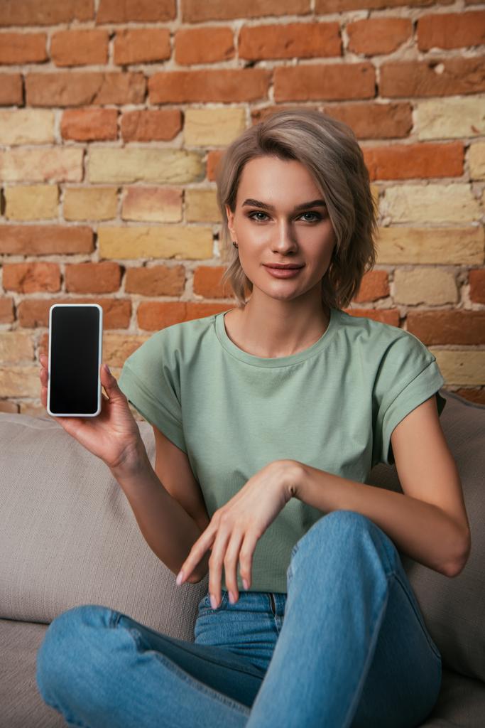 kaunis nuori nainen istuu sohvalla lähellä tiiliseinää ja näyttää älypuhelimen tyhjällä näytöllä
 - Valokuva, kuva