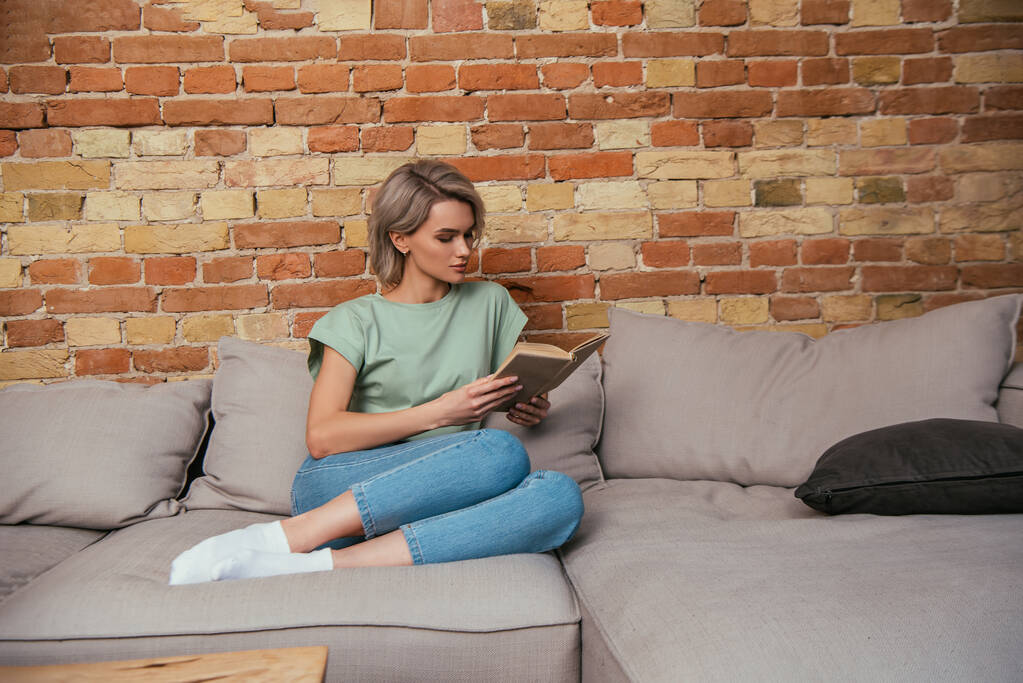 concentré jeune femme lecture livre tandis que assis sur canapé près mur de briques
 - Photo, image