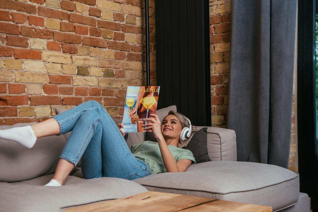 家でソファに横になりながら美とスタイルの雑誌を読んでいる無線ヘッドフォンの笑顔の女性 - 写真・画像