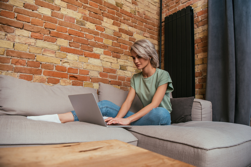 ελκυστική νεαρή γυναίκα που χρησιμοποιεί φορητό υπολογιστή, ενώ στηρίζεται σε καναπέ κοντά σε τοίχο από τούβλα στο σπίτι - Φωτογραφία, εικόνα