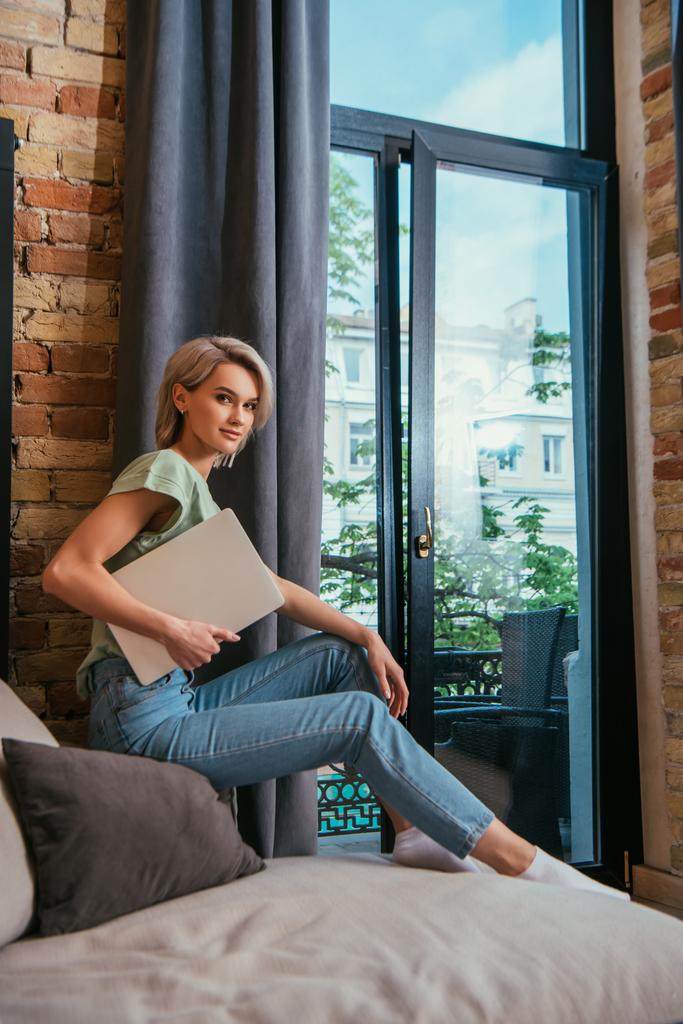 lächelnd blickt eine junge Frau in die Kamera, während sie auf einem Sofa am Fenster sitzt und Laptop in der Hand hält - Foto, Bild