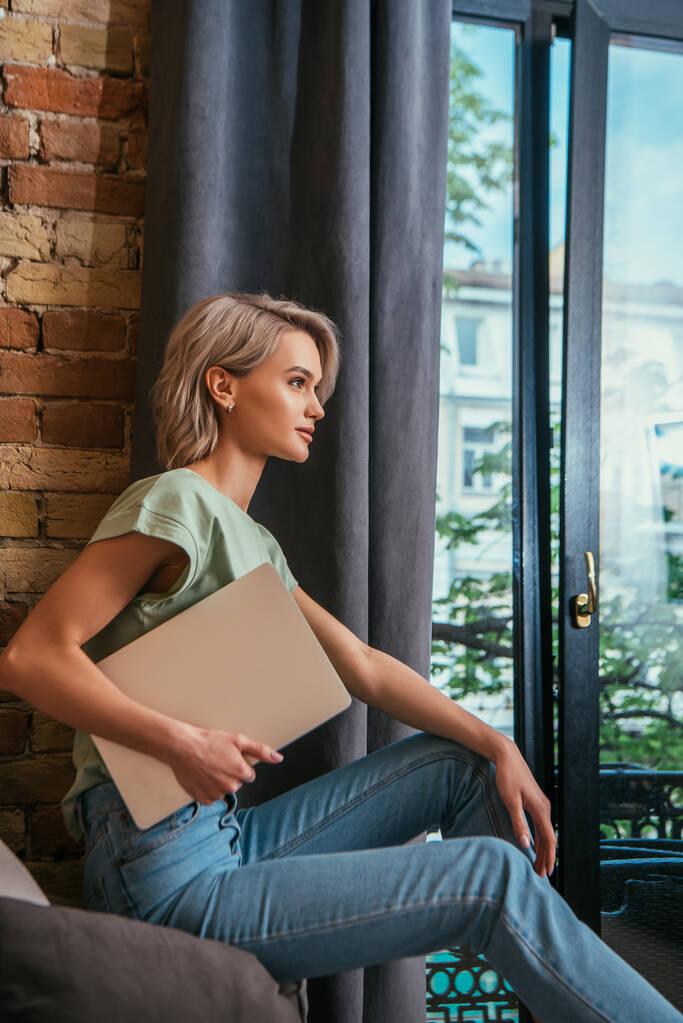 Seitenansicht einer nachdenklichen Frau, die zu Hause am Fenster sitzt und ihren Laptop in der Hand hält - Foto, Bild