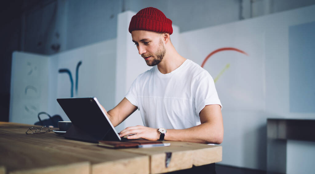 Tartalom professzionális alkalmi hipster felnőtt munkavállaló piros beany és póló ül fa íróasztal nézi notebook képernyő a modern irodában - Fotó, kép