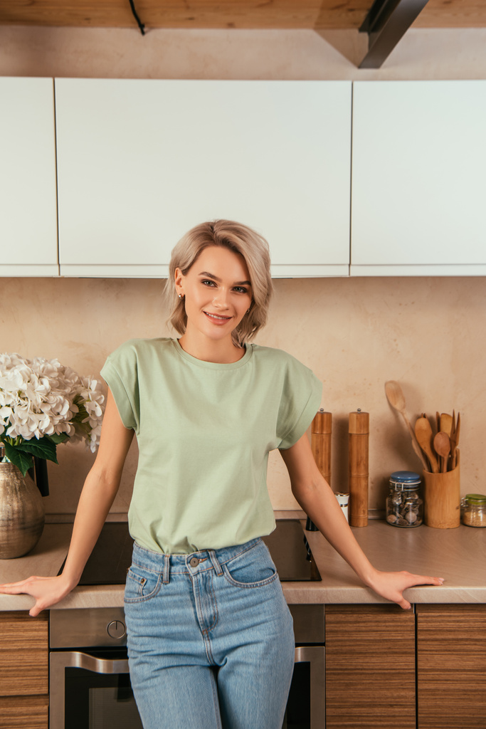 gelukkig, aantrekkelijke vrouw glimlachen op camera terwijl staan in de keuken - Foto, afbeelding