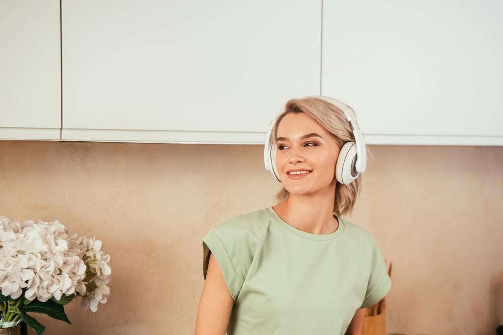 mujer joven sonriente escuchando música en los auriculares y mirando hacia otro lado en la cocina
 - Foto, Imagen