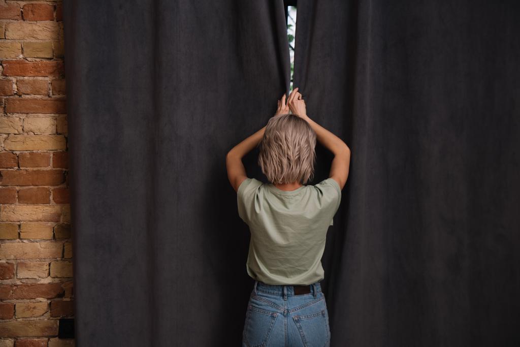 visão traseira da jovem mulher abrindo cortinas escuras na janela em casa
 - Foto, Imagem