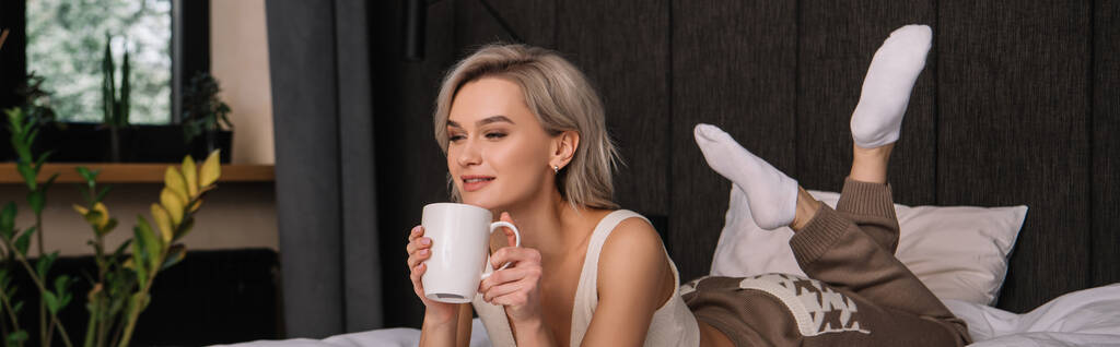 πανοραμική λήψη ελκυστική χαμογελαστή γυναίκα κρατώντας φλιτζάνι τσάι, ενώ χαλαρώνοντας στο κρεβάτι - Φωτογραφία, εικόνα
