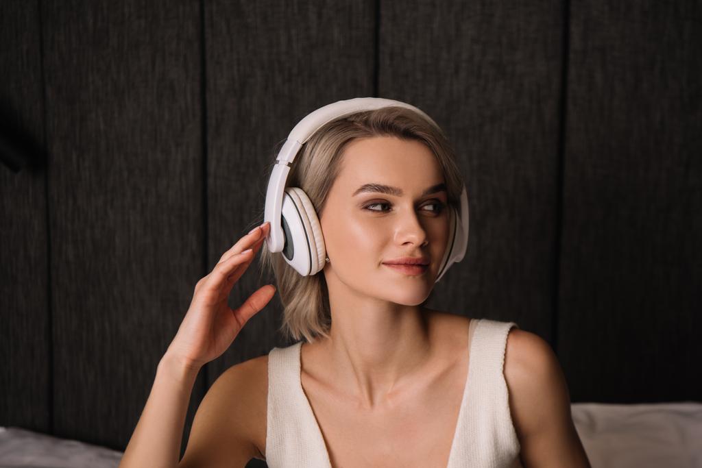 mujer atractiva tocando auriculares inalámbricos mientras mira hacia otro lado
 - Foto, Imagen