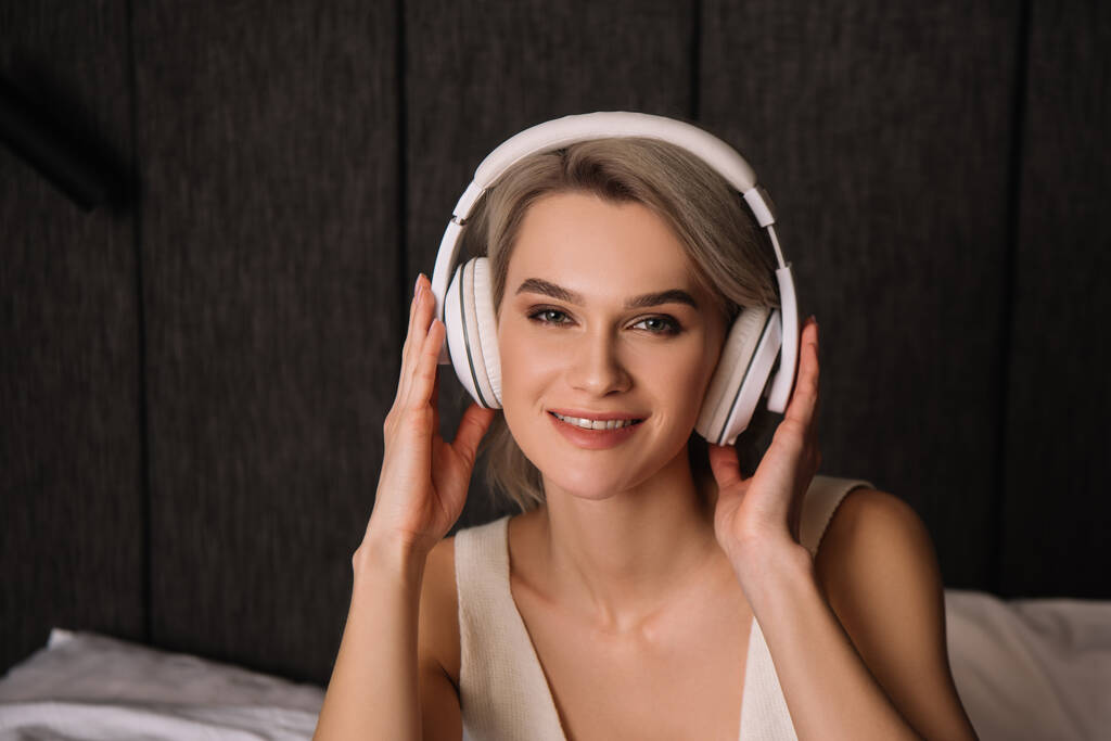 smiling woman looking at camera while touching wireless headphones - Photo, Image