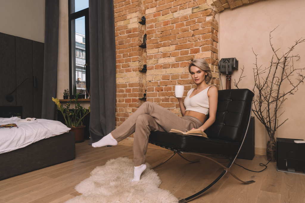 atractiva mujer sosteniendo taza de té y mirando a la cámara mientras está sentado en el sillón cerca de libro en casa
 - Foto, imagen