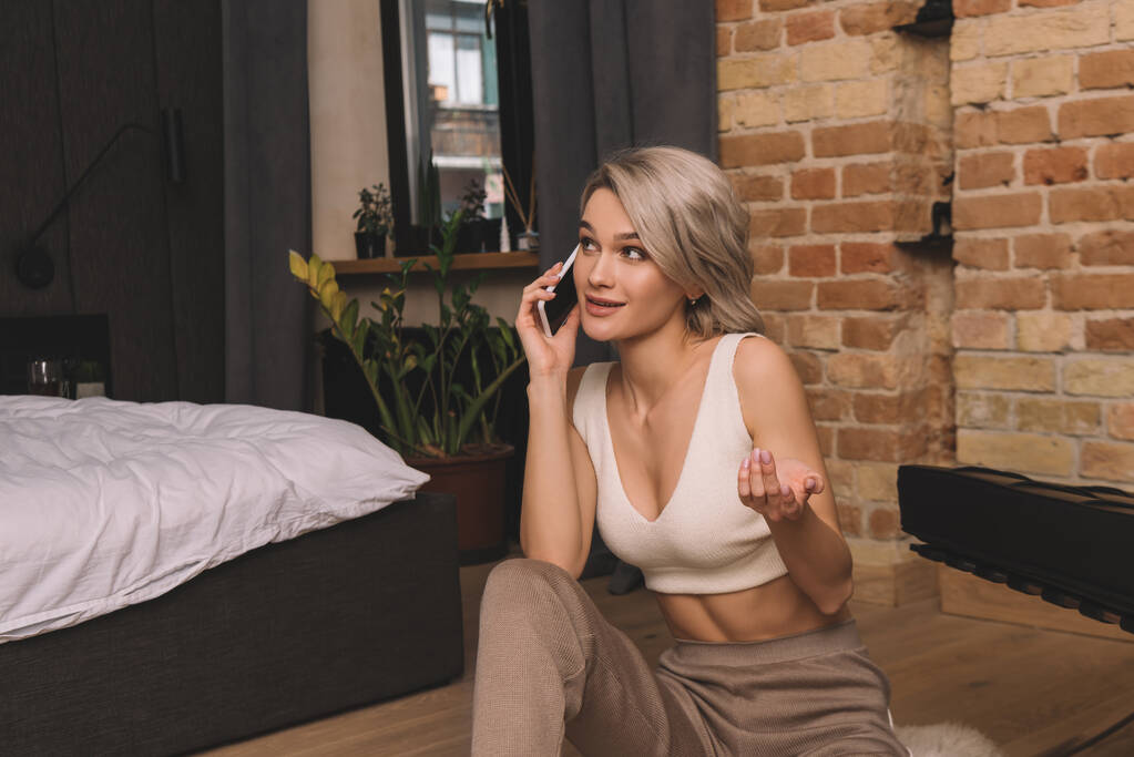 usmívající se, atraktivní žena mluví na smartphone, zatímco sedí na podlaze v ložnici - Fotografie, Obrázek