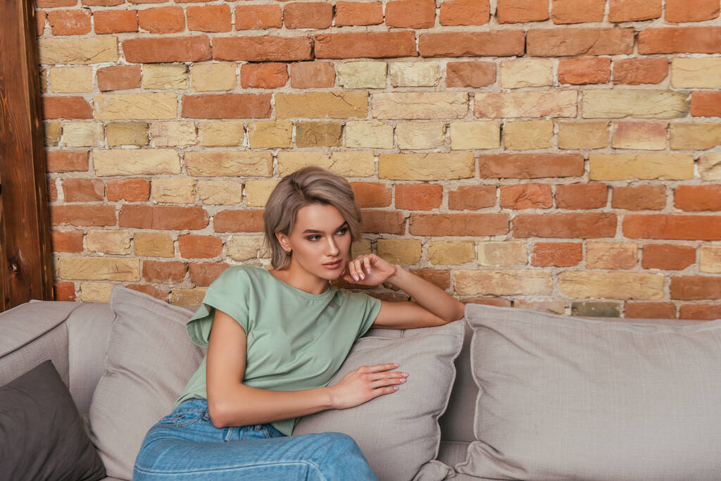 hübsche, verträumte Frau sitzt auf Sofa neben Ziegelwand und schaut weg - Foto, Bild