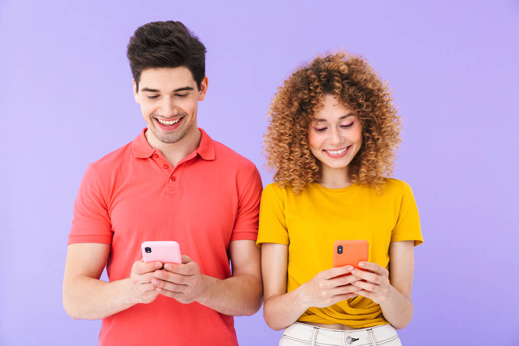 Retrato de personas optimistas caucásicas hombre y mujer en ropa básica sonriendo mientras usan teléfonos celulares juntos aislados sobre fondo violeta
 - Foto, Imagen