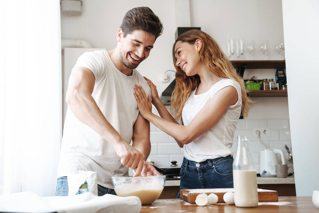 Imagen de pareja adorable regocijándose mientras cocina el desayuno y mezcla de masa en la cocina en casa
 - Foto, imagen