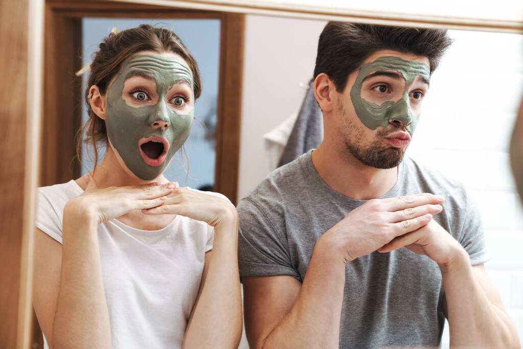 Εικόνα του ευτυχισμένου ζευγαριού άντρας και γυναίκα χαμογελώντας ενώ στέκεται στο μπάνιο με μάσκα προσώπου - Φωτογραφία, εικόνα