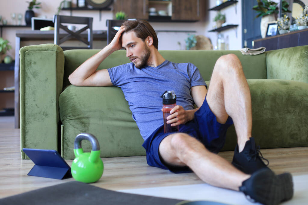 Sportovní muž odpočívá, má přestávku na pitnou vodu po cvičení na podlaze v obýváku. Fintess doma během vlastní izolace - Fotografie, Obrázek