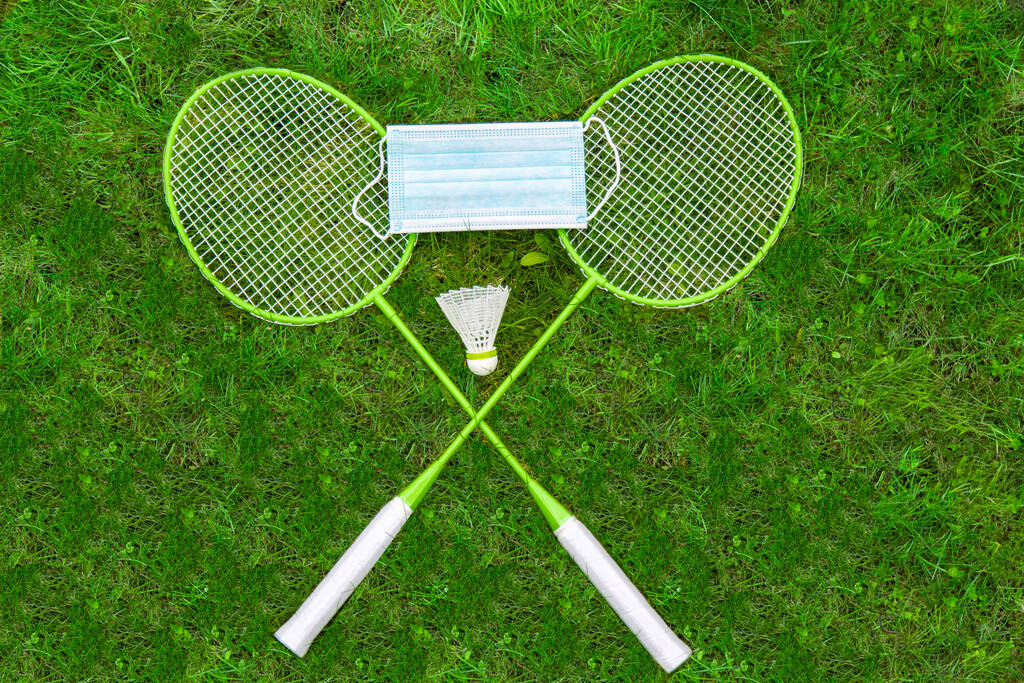 Coronavirus koruma sembolü. Badminton raketleri ve yeşil çimlerde uçan horozlar. Tıbbi maske. Yukarıdan görünüm - Fotoğraf, Görsel