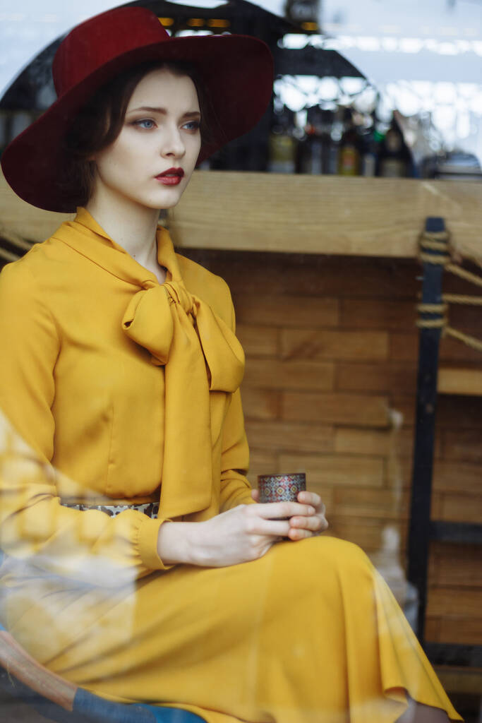 lány egy kávézóban egy csésze kávéval és egy kalapkal.portré érzéki fiatal lány viselt floppy kalap és blúz íj. Gyönyörű barna nő a kávézóban kezében csésze kávét - Fotó, kép