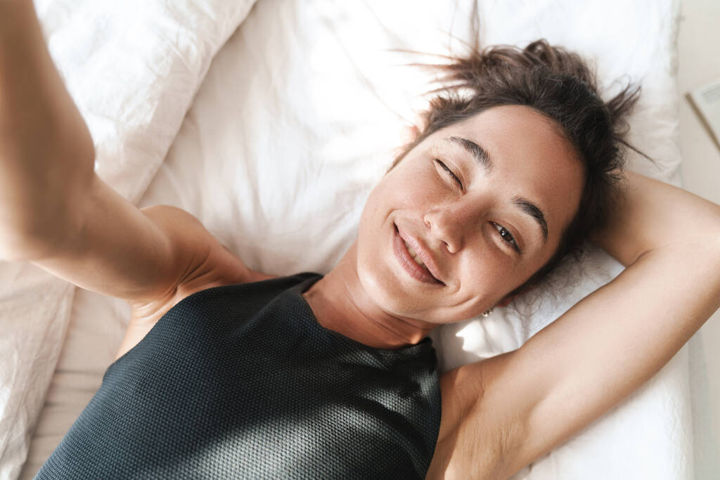 Obrázek potěšené mladé roztomilé ženy doma v posteli vzít selfie fotoaparátem. - Fotografie, Obrázek